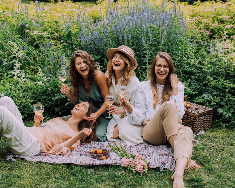 Four women are in a park, they are laughing, sharing a light moment and seem like they are on a picnic or a bestie break