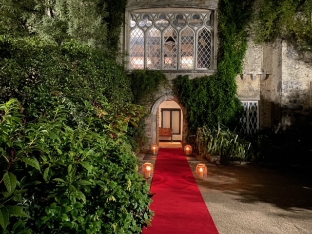You can the outside of the castle and its door in a close up view. It is nighttime and there are lanterns placed leading up to the door and there's a red carpet too