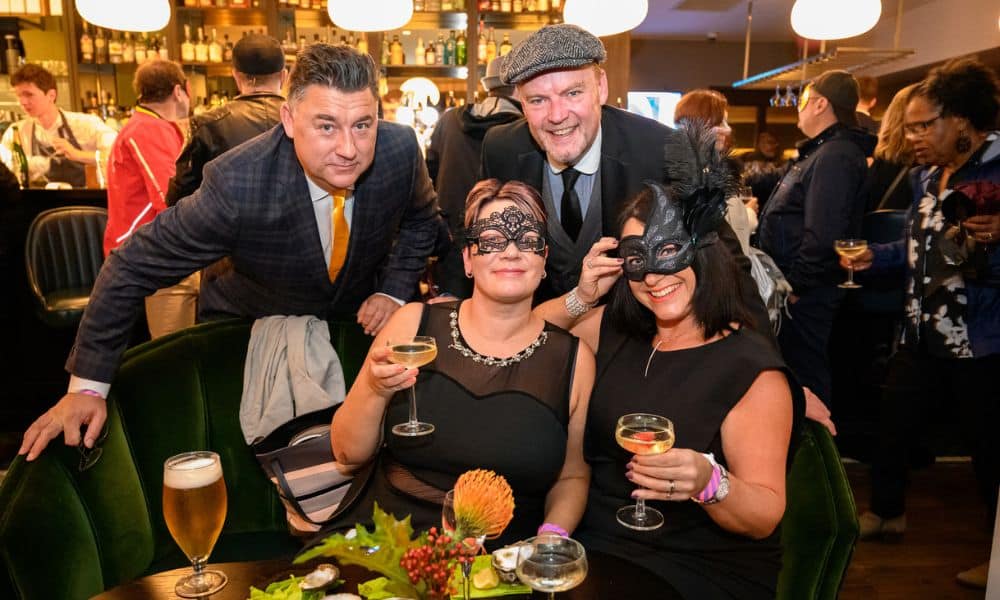 You can see a busy restaurant setting. Women in black dresses and festive eye masks holding up mocktails and men in suits standing behind them
