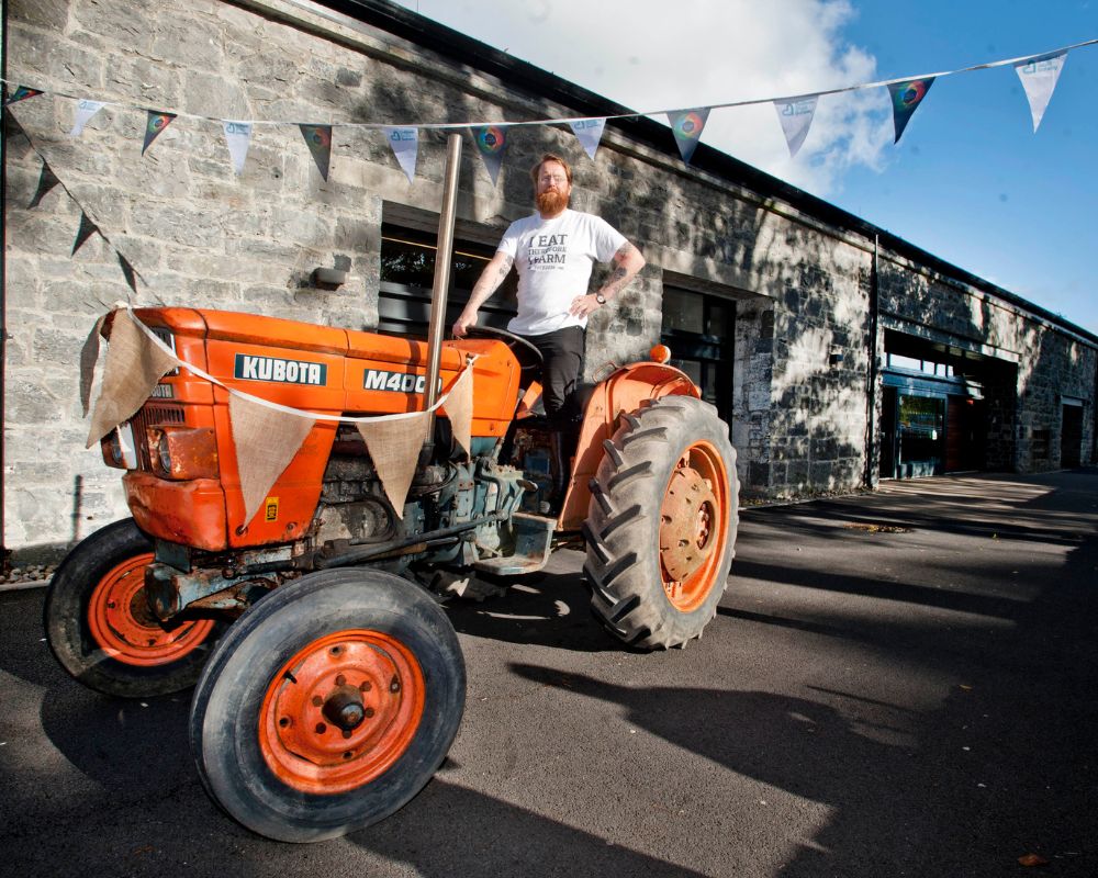 P Mc Mahon, #FOTE2018 director is on an orange tractor and is wearing a white T shirt with the following words 'I eat Therefore I farm' and there is buntings in the framework