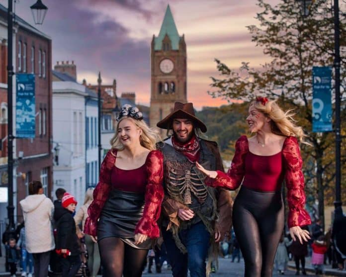 You can see three people - two women and a man dressed up in dark red and black matching Halloween costumes and smiling and walking down the streets of Derry