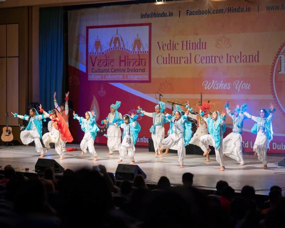 Photograph shows children dressed in white and blue traditional outfits and dancing to a song - a choreographed dance performance for Diwali