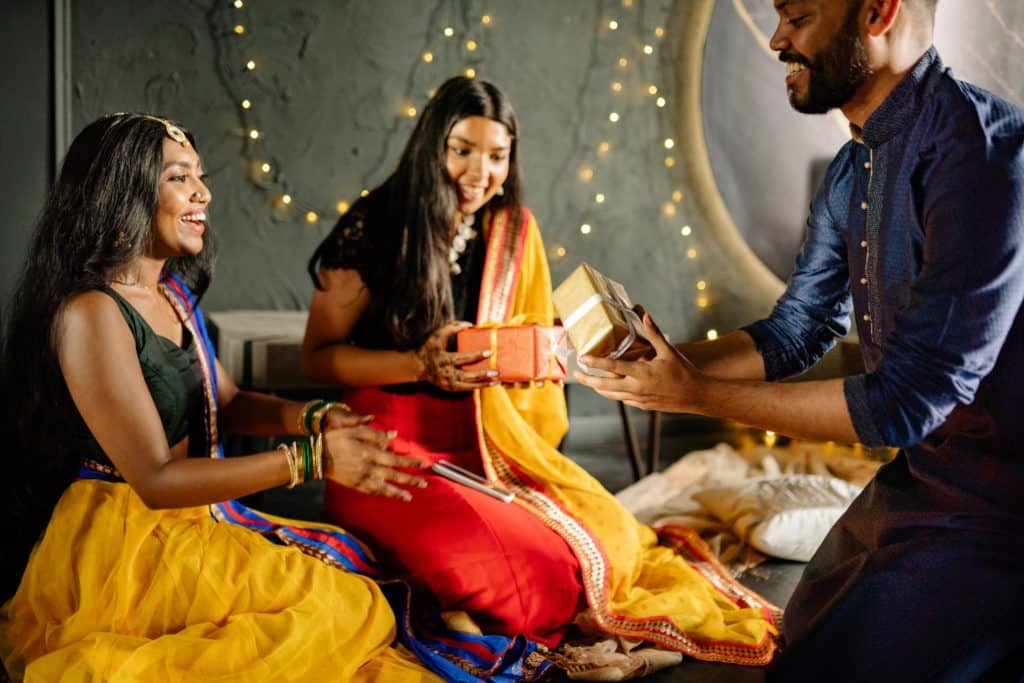 From left to right - photographs show various aspects of Diwali. A young girl holds a platter of sweets, there is a rangoli pattern made up of earthen lamps and colours in the second, in the third two women pray to the Goddess of wealth, in the fourth, a man gives Diwali gifts to family members. 