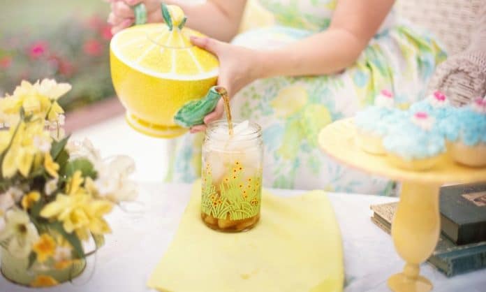 Woman in a floral dress (you can see her hands and not the face) pouring out tea from a yellow melon themed tea pot - she is enjoying afternoon tea with treats